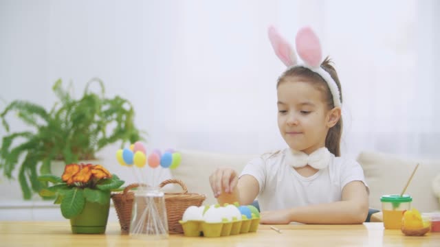 Little-cute-and-adorable-girl-is-smiling-sincerely.-She-takes-an-Easter-egg-from-the-basket-and-looking-at-it.-Girl-is-waving-with-her-liitle-flat-of-hand.-Concept-Easter-holiday.