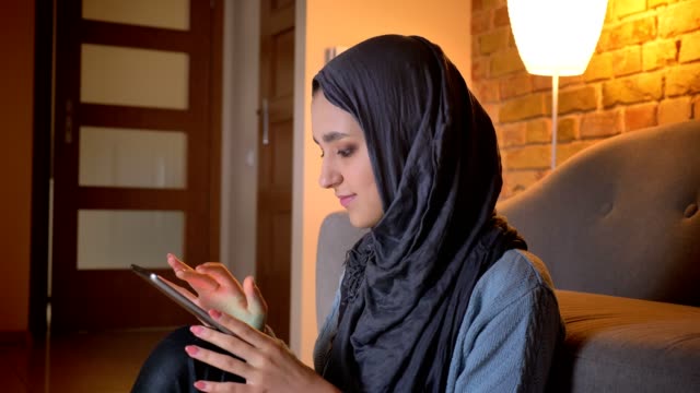 Tiro-de-primer-plano-de-joven-atractiva-mujer-musulmana-en-hijab-escribiendo-en-el-teléfono-mientras-está-sentado-en-el-suelo-en-la-puerta-de-casa-acogedora