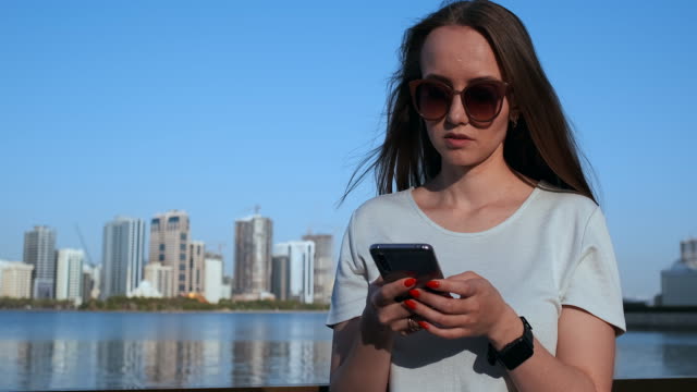 Beautiful-girl-with-long-hair-in-sunglasses-using-smartphone-app-at-sunset-river-quay-near-4K.