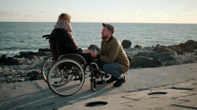 Man-is-holding-hands-of-sick-young-woman-in-wheelchair-and-talking-with-her