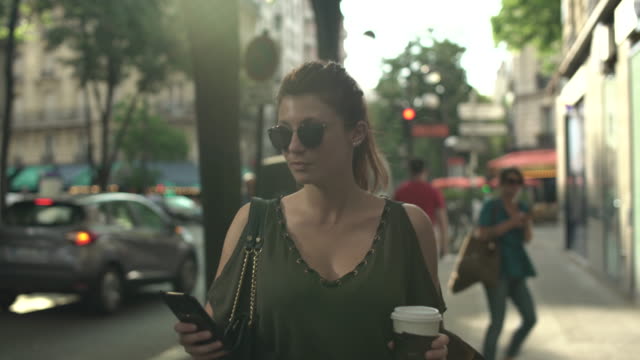 Attraktive-kaukasische-Frau-mit-Sonnenbrille,-Sommersprossen,-Piercings-und-roten-Haaren-scrollen-Social-Media-auf-ihrem-Smartphone-zu-Fuß-durch-die-Straße,-während-sonnigen-Sommer-in-Paris.-4K-UHD.-supermodern.