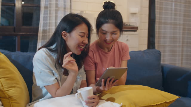 Lesbian-lgbt-women-couple-using-tablet-at-home,-Asian-female-happy-using-technology-check-social-media-in-internet-together-while-lying-sofa-in-living-room-in-night.-Lover-celebrate-holiday-concept.