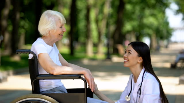 Smiling-nurse-talking-with-disabled-elderly-woman-in-park,-rehabilitation-center