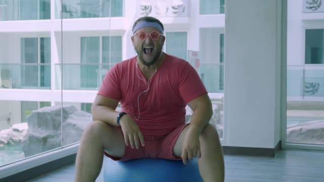 Funny-fat-male-in-pink-glasses-and-in-a-pink-t-shirt-is-engaged-on-a-fit-ball-in-the-gym-depicting-a-girl.-A-wonderful-man-listens-to-music-and-dances-on-the-ball-in-the-gym