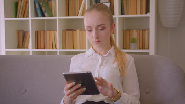 Nahaufnahme-Porträt-der-jungen-hübschen-kaukasischen-blonden-Studentin-mit-dem-Tablet,-das-auf-die-Kamera-schaut,-die-auf-der-Couch-drinnen-in-der-Wohnung-sitzt