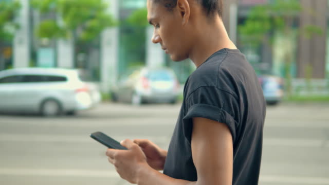 Profil-afrikanischen-amerikanischen-Mann-zu-Fuß-halten-Smartphone-entlang-der-Straße-in-der-Stadt