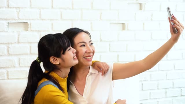 Happy-Asian-teenage-daughter-and-middle-aged-mother-taking-selfie-or-video-calling