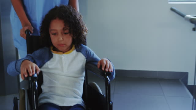 Vista-frontal-del-doctor-empujando-a-un-niño-afroamericano-en-silla-de-ruedas-en-el-pasillo-del-hospital-4k