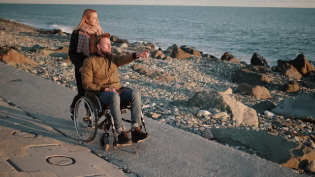 Young-woman-is-nursing-sick-disabled-man,-rolling-stroller-on-sea-shore