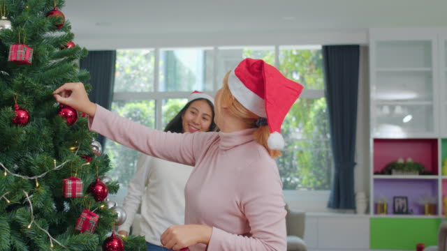 Asian-women-friends-decorate-Christmas-tree-at-Christmas-festival.-Female-teen-happy-smiling-celebrate-xmas-winter-holidays-together-in-living-room-at-home.-Slow-motion-shot.