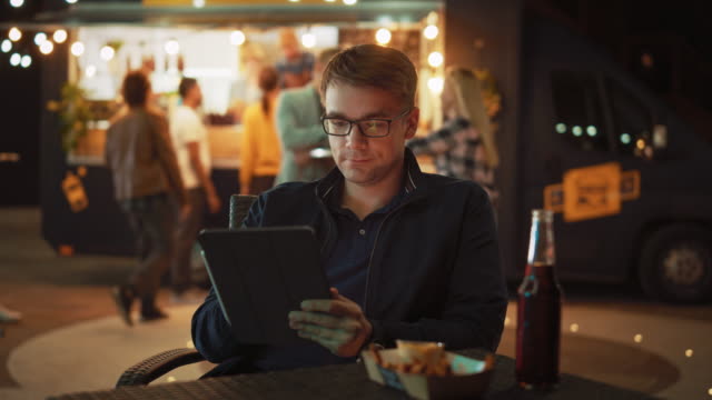 Der-hübsche-junge-Mann-in-der-Brille-benutzt-ein-Tablet,-während-er-an-einem-Tisch-in-einem-Outdoor-Street-Food-Cafe-sitzt-und-Fries-isst.-Er-surft-im-Internet-oder-in-sozialen-Medien-und-veröffentlicht-ein-Status-Update.