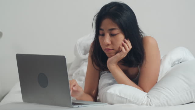Junge-asiatische-Frau-mit-Laptop-Überprüfen-Social-Media-Gefühl-glücklich-lächeln,-während-auf-dem-Bett-liegen-duponieren,-nachdem-sie-morgens-im-Haus-aufwachen,-attraktive-thai-Mädchen-lächelnd-entspannen-im-Schlafzimmer-zu-Hause-Konzept.