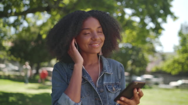 Eine-afrikanische-amerikanische-junge-Frau,-die-im-Park-steht-und-Musik-auf-Kopfhörern-auf-ihrem-Handy-hört---lächelnd-und-tanzende-Frau-im-Park