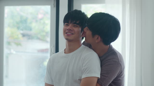 Asiatische-Homosexuell-Paar-stehen-und-umarmen-in-der-Nähe-des-Fensters-zu-Hause.-Junge-asiatische-LGBTQ+-Männer-küssen-glückliche-Entspannen-Ruhe-zusammen-verbringen-romantische-Zeit-im-Wohnzimmer-im-modernen-Haus-im-Morgenkonzept.