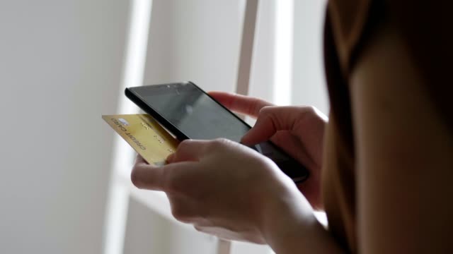 Schließen-Sie-die-Hände-einer-schönen-asiatischen-kaukasischen-Frau-mit-Smartphone-Shopping-online-und-verwenden-Sie-die-Kreditkartenzahlung,-während-neben-dem-Fenster-im-Büroraum-stehen.