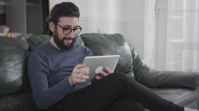 Young-Caucasian-man-with-black-hair-and-beard-wearing-eyeglasses-using-tablet-as-sitting-at-the-couch.-Intelligent-guy-using-social-media.-Internet-addiction,-facebook,-instagram.