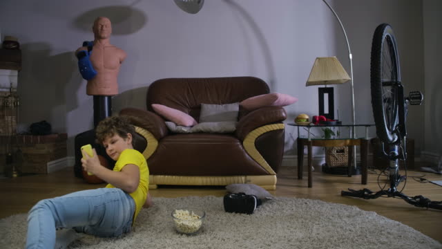 Timelapse-of-curly-haired-child-in-casual-clothes-talking-on-smartphone-and-eating-popcorn.-Caucasian-boy-sitting-on-the-floor,-them-moving-on-the-couch.-Leisure,-resting.-Cinema-4k-ProRes-HQ.