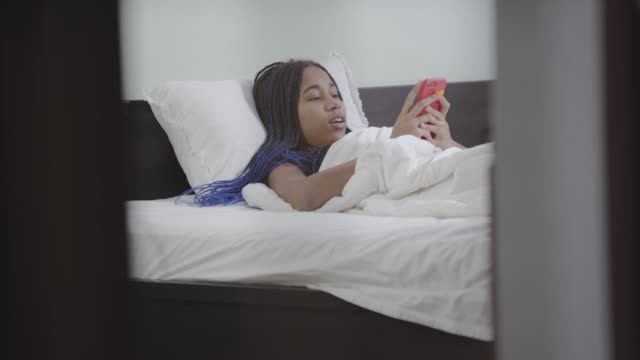Porträt-von-nachdenklichen-Mädchen,-die-Smartphone,-Tippen-und-Lächeln.-Afroamerikanische-Teenager-ruht-im-weißen-Bett-zu-Hause.-Social-Media,-Internetsucht,-Lebensstil.