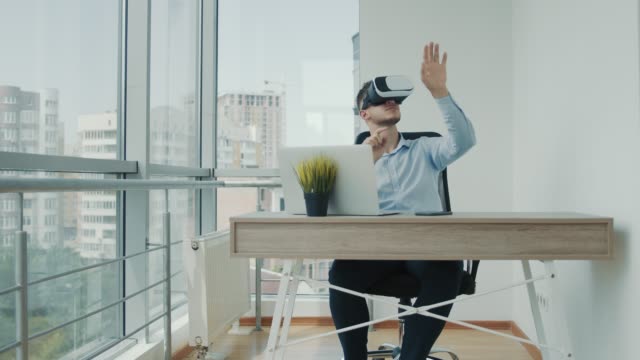 A-young-man-sitting-at-a-desk-in-the-office-uses-augmented-reality-glasses-to-work-on-business-projects-in-various-fields.