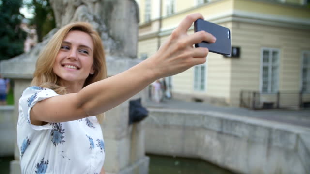 Young-girl-taking-selfie-with-smartphone-sitting-on-fountain-in-the-city.-Happy-young-woman-smiling-posing-for-self-portrait-using-iPhone-smart-phone-camera.-Tourism,-instagram,-modern-technology.