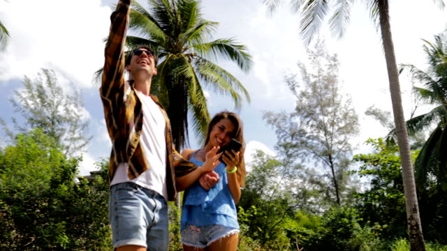 Couple-Walk-Using-Cell-Smart-Phone-Under-Palm-Trees,-Man-Hold-Hand-Up-Video-Call,-Smiling-Man-And-Woman-Online-Communication