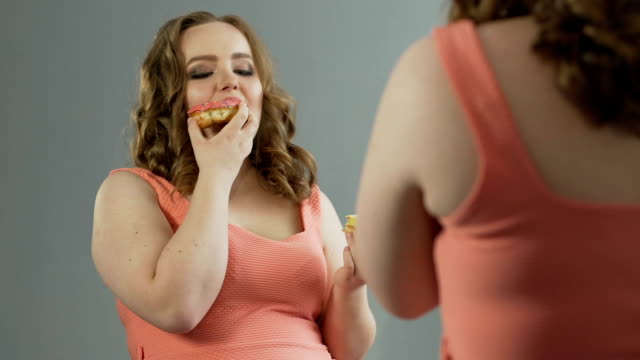 Mujer-gordita-disfrutando-de-dos-donuts-delante-espejo,-admirando-su-forma-del-cuerpo
