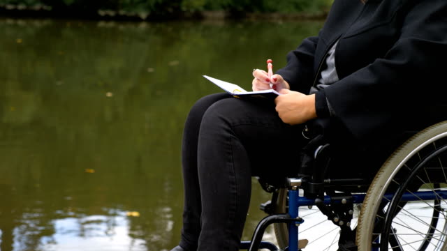 close-up-on-woman-on-wheelchairs-writes-a-love-letter-near-the-lake
