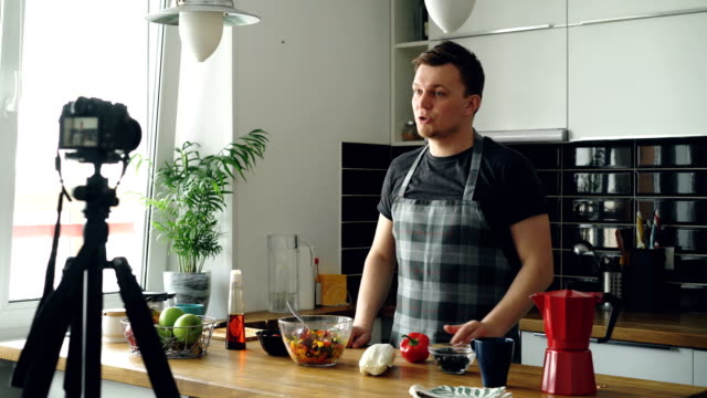 Handsome-guy-recording-video-food-vlog-about-healthy-cooking-on-digital-camera-in-the-kitchen-at-home.-Vlogging-and-social-media-concept