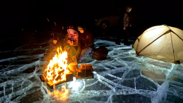 Four-travelers-by-fire-right-on-ice-at-night.-Campground-on-ice.-Tent-stands-next-to-fire.-People-are-warming-around-campfire.-Photographer-in-shoots-on-camera-in-tripod.