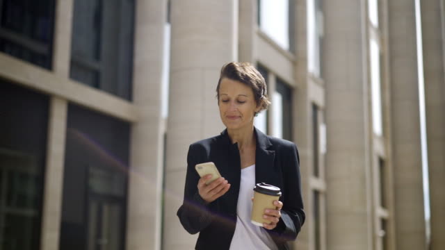 Smiling-middle-aged-woman-with-short-hair-walking-down-street-with-takeaway-coffee-cup-and-text-messaging-on-her-cell-phone-looking-happy-and-inspired