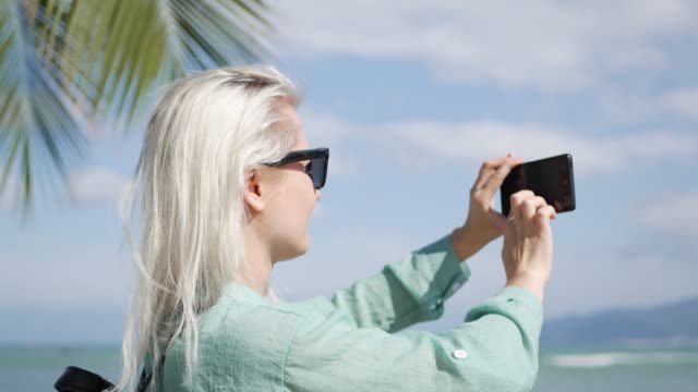 Young-beautiful-slim-woman-with-long-blonde-hair-in-sunglasses-and-green-shirt-standing-near-palm-tree-and-take-a-pictures-by-smartphone-on-a-blue-sky-background.-Girl-using-mobile-phone