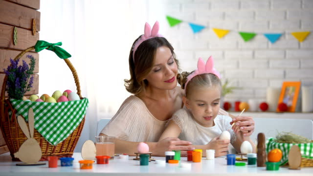 Docente-madre-hija-decorar-huevos,-familia-en-diadema-lindo-sentado-en-la-mesa