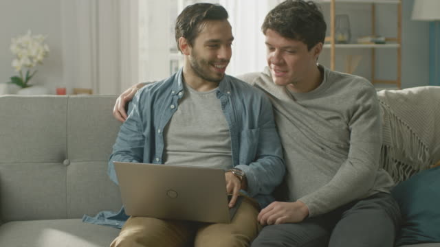 Adorable-Male-Gay-Couple-Spend-Time-at-Home.-They-Sit-on-a-Sofa-and-Use-the-Laptop.-They-Browse-Online.-Partner-Puts-His-Hand-Around-His-Lover.-Room-Has-Modern-Interior.