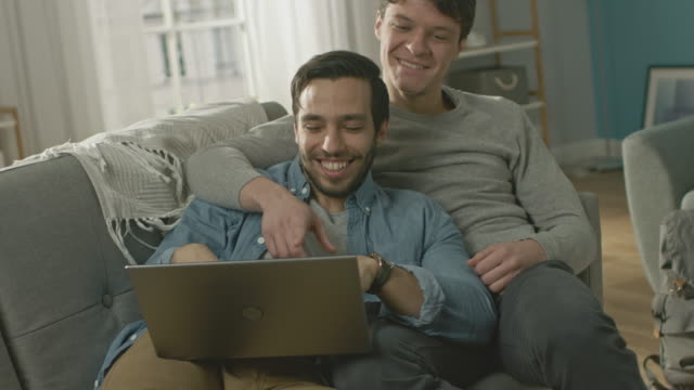 Cute-Male-Gay-Couple-Spend-Time-at-Home.-They-are-Lying-Down-on-a-Sofa-and-Use-the-Laptop.-They-Browse-Online.-Partner's-Hand-is-Around-His-Lover.-They-Smile-and-Laugh.-Room-Has-Modern-Interior.