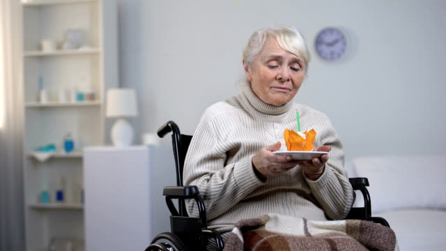 Alte-Frau-im-Rollstuhl-Ausblasen-der-Kerzen-auf-Kuchen-und-Blick-in-die-Kamera-zu-weinen