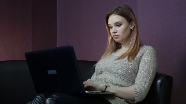 A-young-woman-works-at-a-computer-while-sitting-on-a-small-couch.