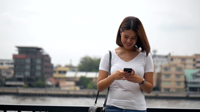 Hermosa-joven-asiática-usando-el-teléfono-inteligente-disfrutando-de-leer-las-redes-sociales-al-lado-de-la-valla-en-el-río.