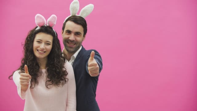 Young-couple-standing-on-a-pink-background.-During-this,-the-gesture-class-shows-a-smile,-looking-at-the-camera.-Happy-family-is-preparing-for-Easter,-with-the-ears-of-a-pink-rabbit-on-its-head.