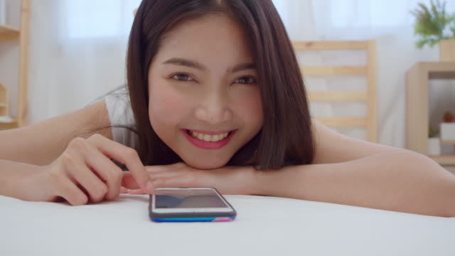 Slow-motion---Young-Asian-woman-using-smartphone-checking-social-media-feeling-happy-smiling-while-lying-on-bed-after-wake-up-in-the-morning,-Attractive-Japanese-girl-smiling-relax-in-bedroom-at-home.