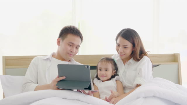 Asiatische-Familie-lacht-und-schaut-in-digitalen-Tablet,-während-auf-dem-Bett-im-Schlafzimmer-liegen