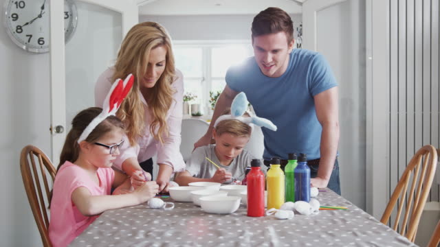 Familia-sentada-alrededor-de-la-mesa-de-la-cocina-con-los-padres-ayudando-a-los-niños-a-pintar-huevos-para-Semana-Santa---tiro-en-cámara-lenta