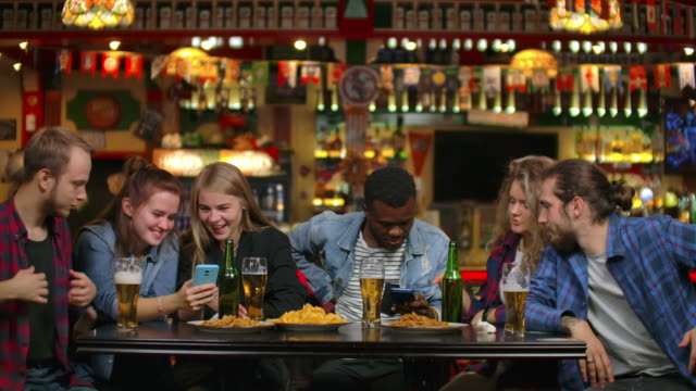 Grupo-de-amigos-de-estudiantes-de-bar-beben-cerveza-y-miran-las-pantallas-de-los-teléfonos-inteligentes-sonriendo