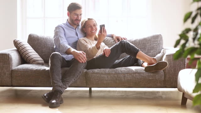 Happy-couple-laughing-holding-phone-talking-relaxing-together-on-sofa
