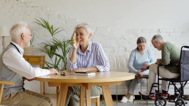Tracking-shot-of-elderly-couple-sitting-at-table-and-enjoying-friendly-talk,-senior-woman-and-disabled-senior-man-in-wheelchair-playing-cards-in-background-in-common-room-of-nursing-home