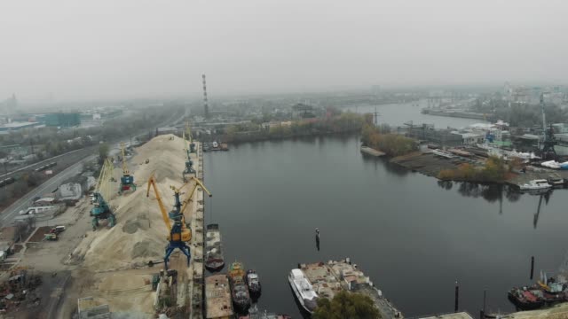 Aerial-view-of-industrial-grey-city-covered-in-fog-and-smog.-Cargo-trains-and-railway-station-near-river-docks-and-cargo-port-cranes