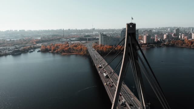 Ukrainische-Flagge-weht-auf-Brücke-verbindet-zwei-Ufer-der-Metropole.-Luft-Drohnenansicht-der-Betonbrücke-mit-viel-Befahrenem-Autoverkehr.-Flug-einer-Drohne-über-Brücke-mit-schönem-Stadtbild-mit-Fluss.-Kiew,-Ukraine