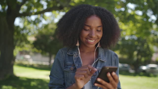 Smiling-portrait-of-an-african-american-young-woman-with-earphones-in-her-ears-enjoying-chatting-and-taking-on-mobile-phone-in-park-smiling