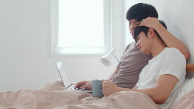 Asian-Gay-men-couple-using-computer-laptop-and-drinking-coffee-at-modern-home.-Young-Asia-lover-male-happy-relax-rest-together-after-wake-up,-watching-movie-lying-on-bed-in-bedroom-at-house-in-morning