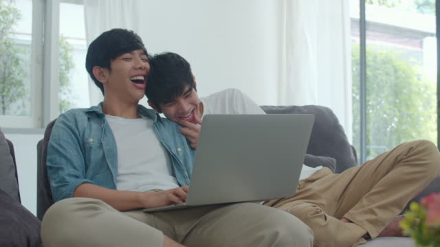 Young-Gay-couple-using-computer-laptop-at-modern-home.-Asian-LGBTQ-men-happy-relax-fun-using-technology-watching-movie-in-internet-together-while-lying-sofa-in-living-room-at-house-concept.