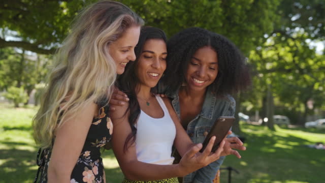 Glückliche-junge-Gruppe-von-multiethnischen-Freundinnen,-die-mit-Smartphone-Technologie-surfen-und-an-einem-sonnigen-Tag-Momente-miteinander-im-Park-teilen